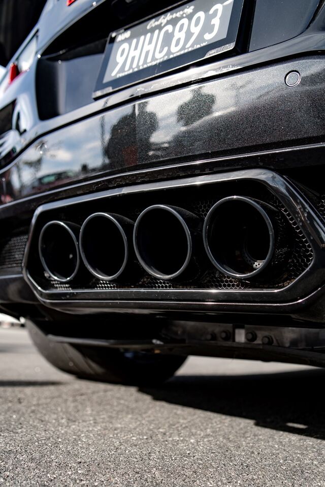 A black car's rear exhaust pipes are prominently displayed, showing four chrome outlets below a license plate.