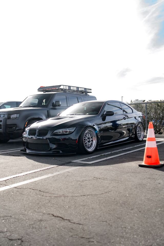 A black BMW M3 sits in a parking lot next to a black SUV with a roof rack.