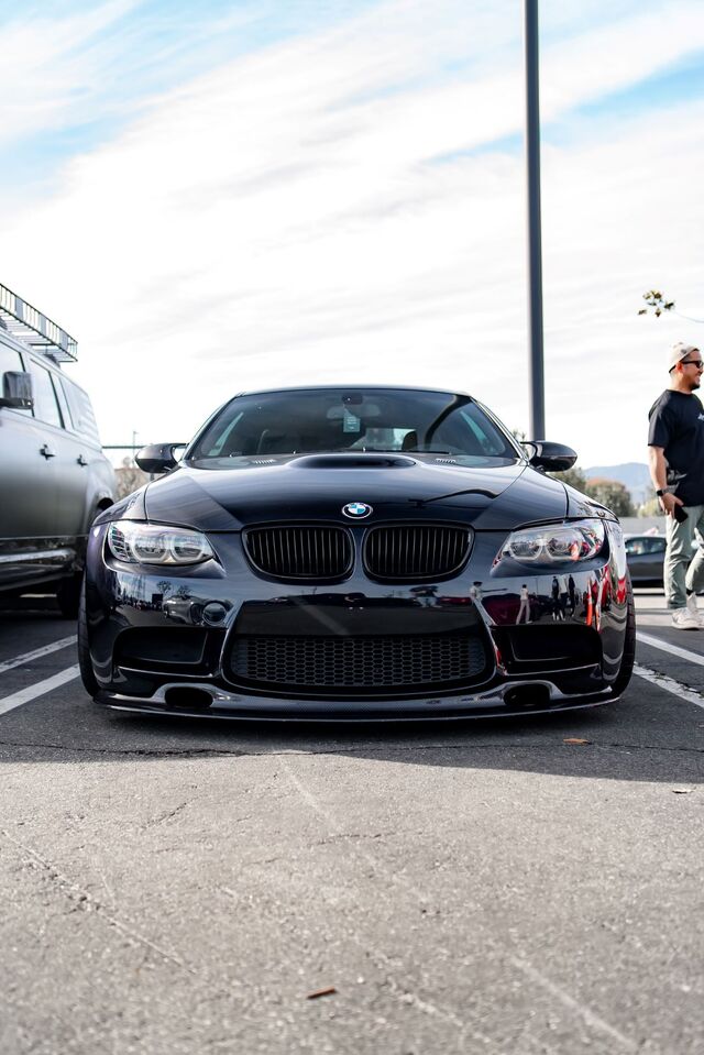 A sleek, black BMW coupe sits parked on pavement with a blurred background of people and vehicles.