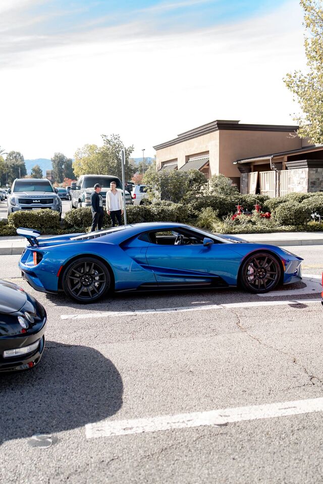 A bright blue Ford GT sits in a parking lot next to a black car and two people standing nearby.