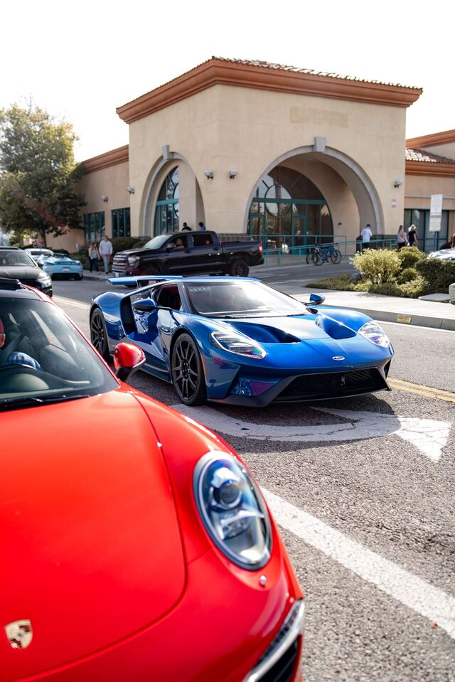 A blue Ford GT and a red Porsche are parked at a car show near a brick building.