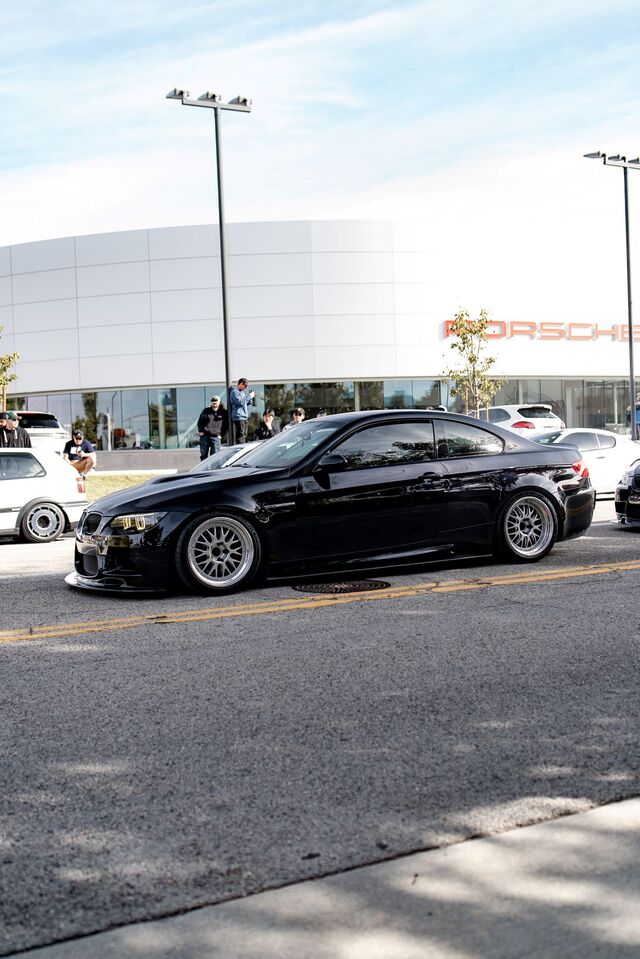 A sleek black BMW sedan sits parked in a car meet outside a Porsche dealership.