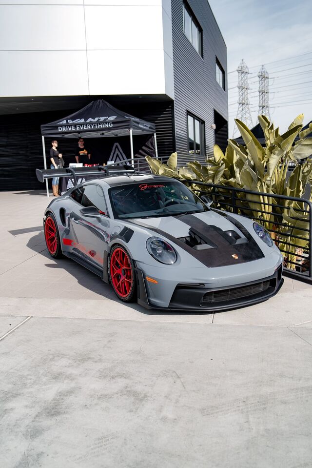 A gray Porsche 911 with red accents sits parked in front of a building and pop-up tent.