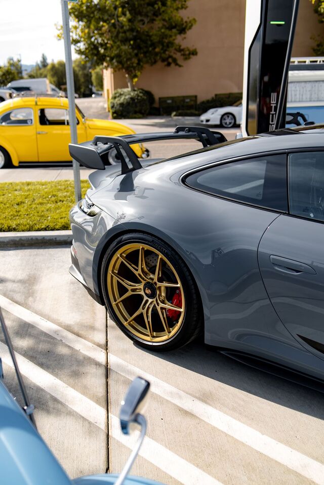 A gray Porsche with gold wheels and a large spoiler is parked in a lot.
