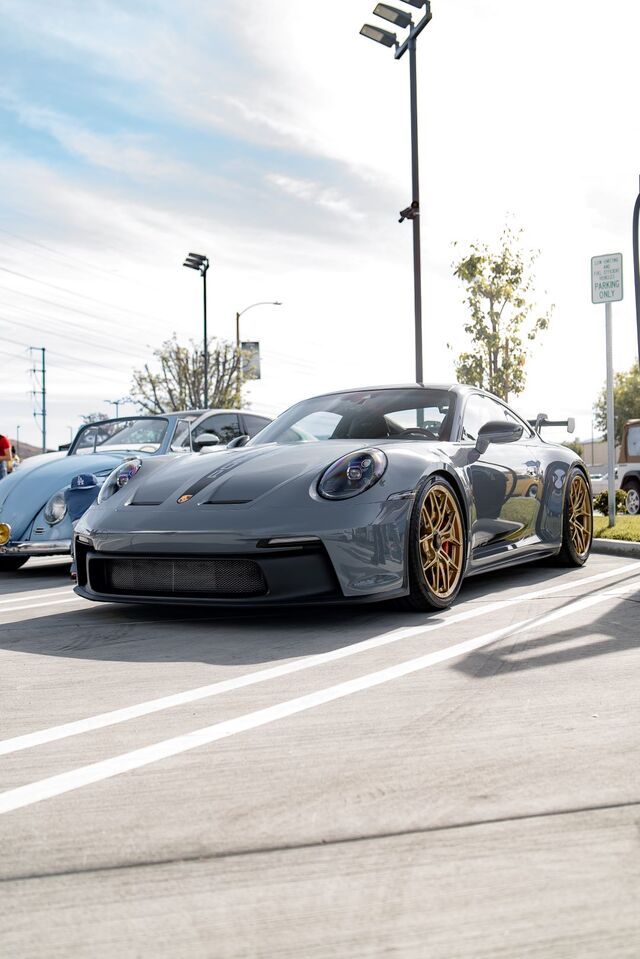 A gray Porsche 911 with gold wheels sits parked in a lot with a street sign visible nearby.
