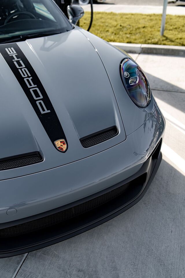 A gray Porsche with a black racing stripe and prominent headlights sits parked on pavement.