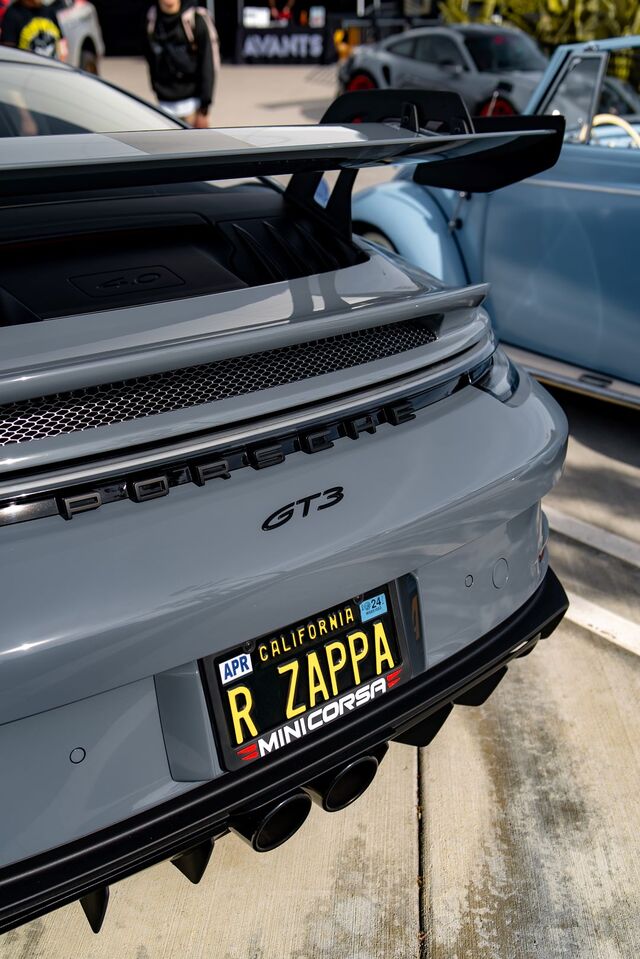 A gray Porsche GT3 with a large rear wing sits parked, displaying a California license plate.