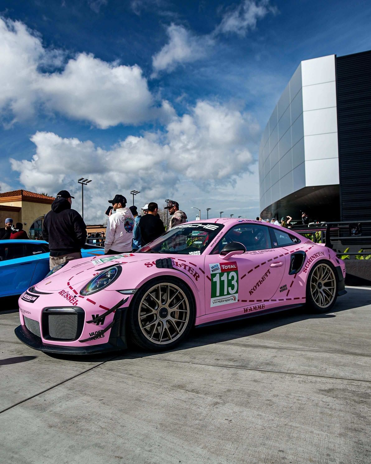 A pink Porsche GT2RS with racing decals and the number 113 on its side, at an outdoor car show.