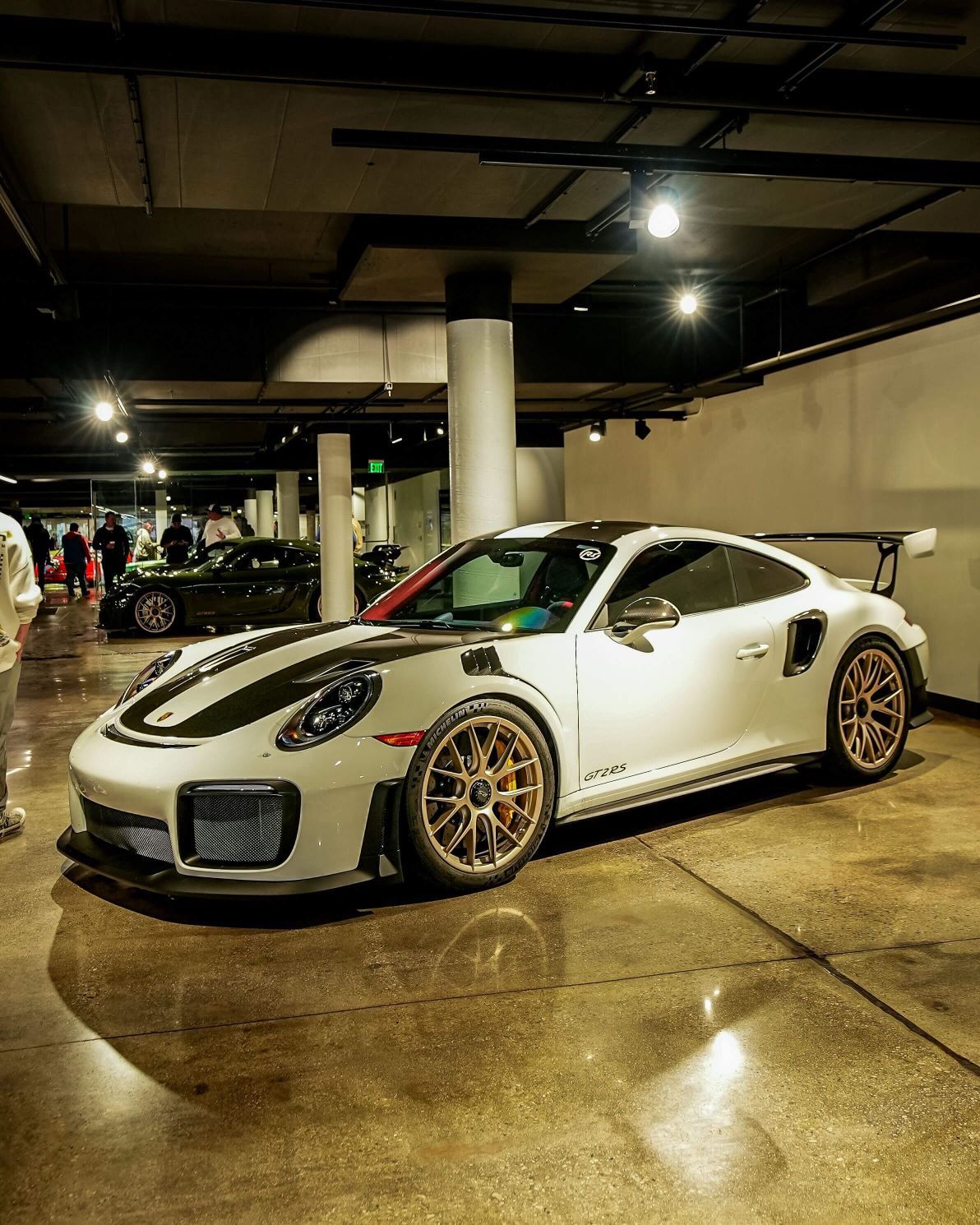 White Porsche 911 GT2 RS with black racing stripes and bronze wheels, parked indoors on a concrete floor.