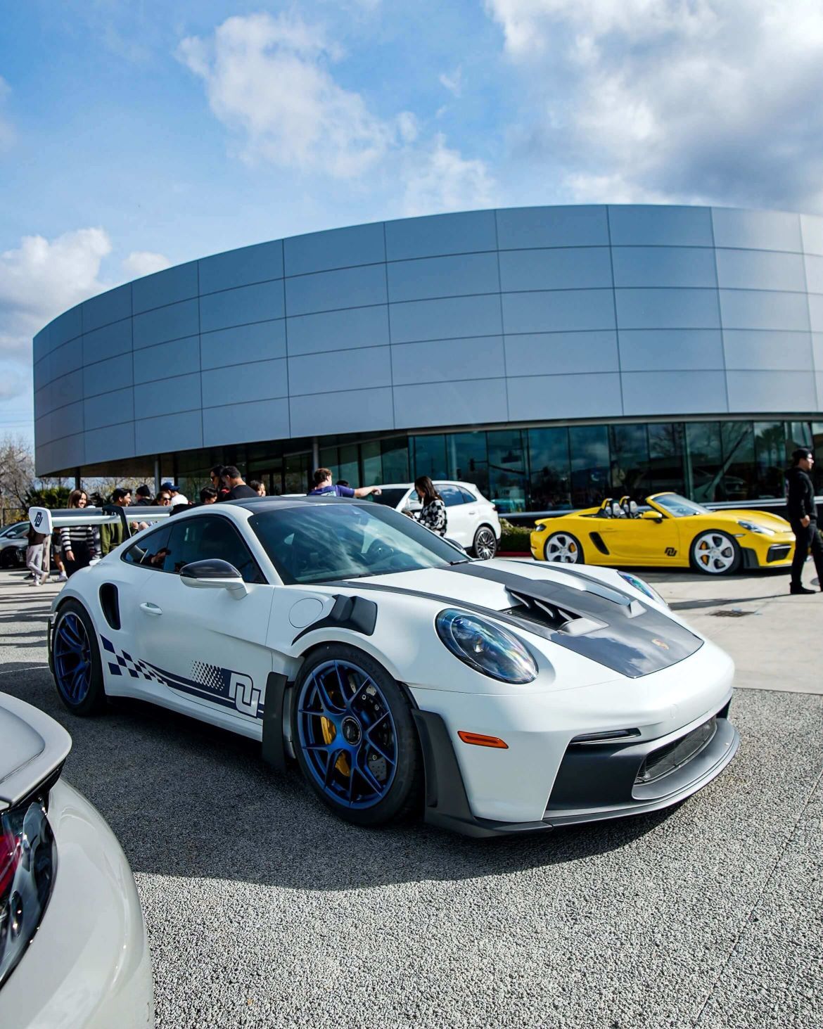 White Porsche GT3 RS with blue wheels, black hood stripes, and a checkered decal, parked at an event.