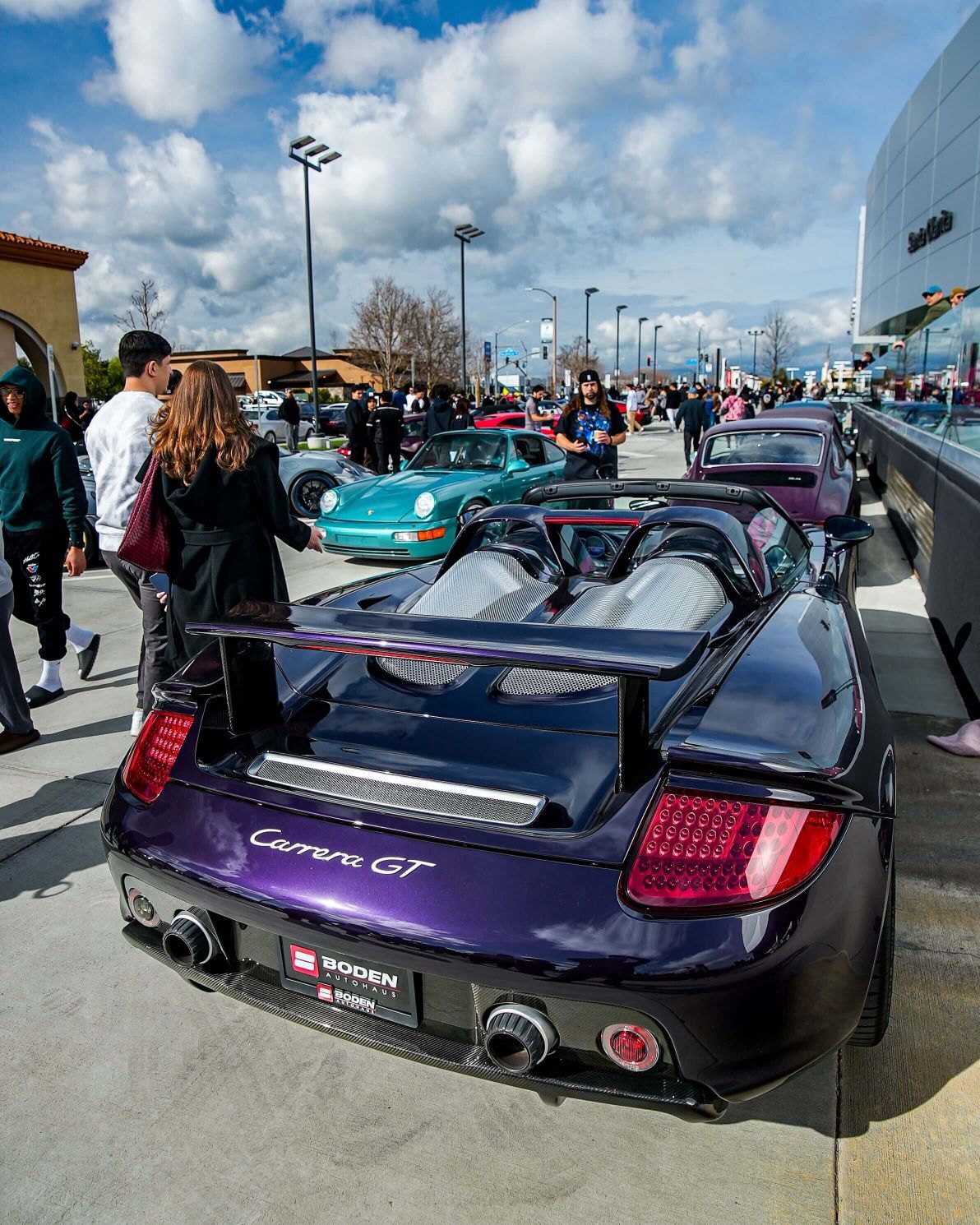 A glossy, deep purple Porsche Carrera GT with its rear visible, parked amidst a crowd at an outdoor car show.