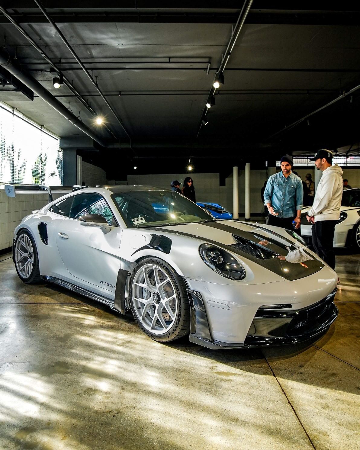 A light grey Porsche GT3 RS with a black hood stripe, parked indoors with two men talking in the background.