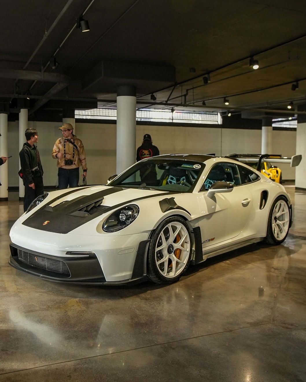 White Porsche 911 GT3 with black stripes on hood in a showroom with people and another car in background.