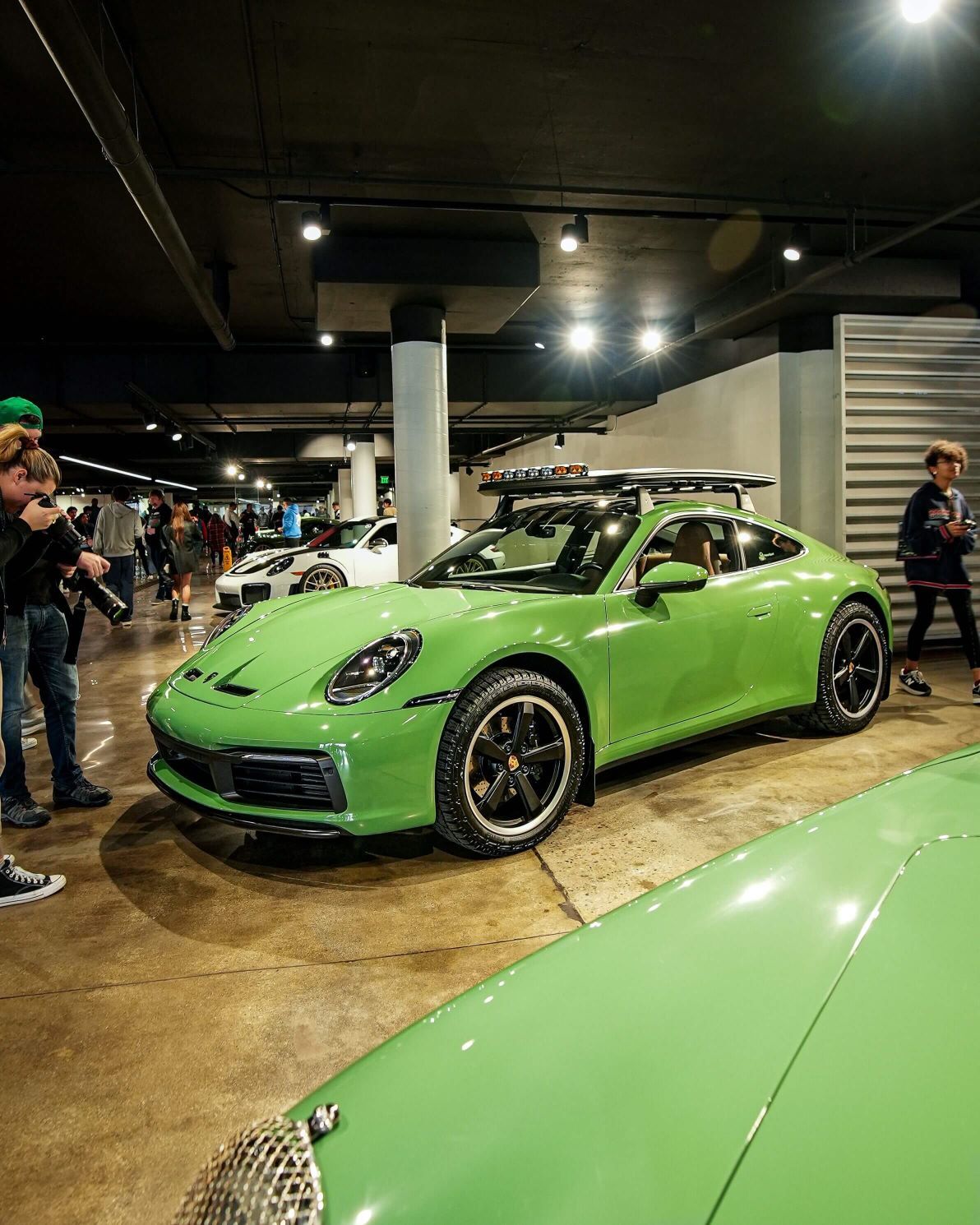 Lime green Porsche 911 with a roof rack and surfboard, in a brightly lit exhibition hall with people around.