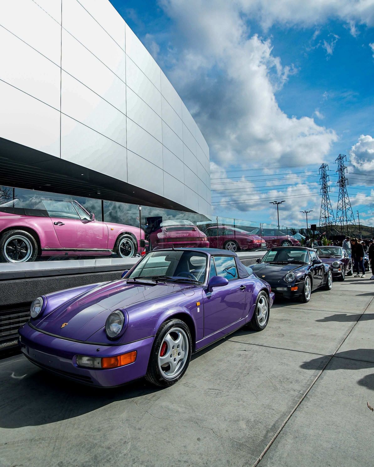 A lineup of classic Porsches, with a purple convertible in the foreground, next to a modern white building.