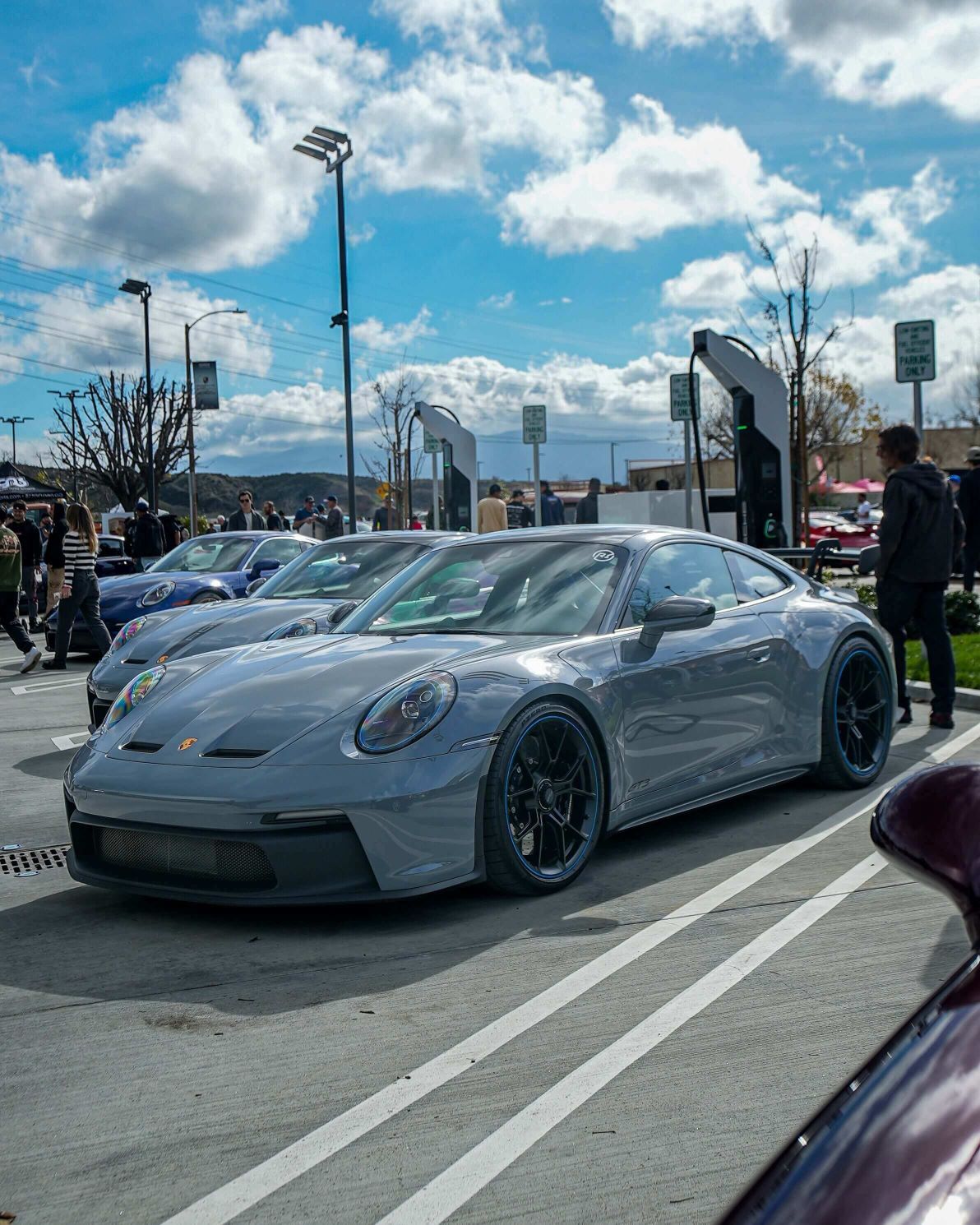 A gray Porsche 911 GT3 parked in an outdoor lot on a sunny day with people and electric charging stations.