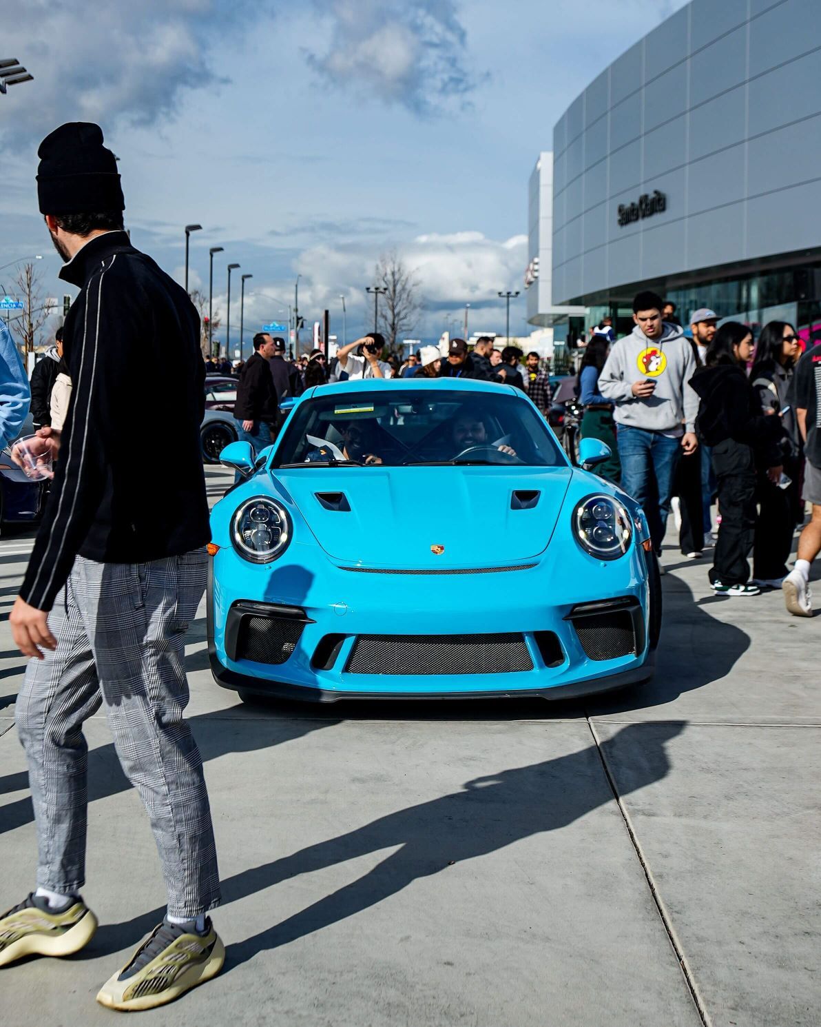 A bright blue Porsche GT3 surrounded by a crowd of people under a cloudy sky.