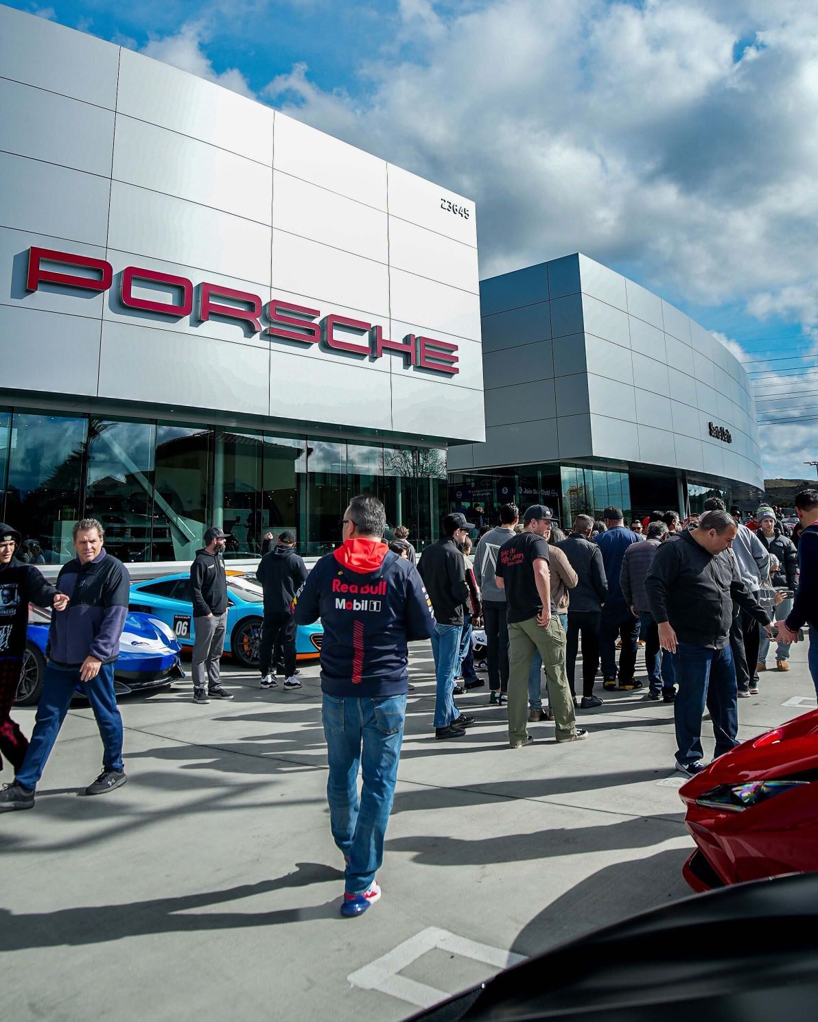 Red Porsche dealership with a crowd of people and luxury cars parked outside under a partly cloudy sky.