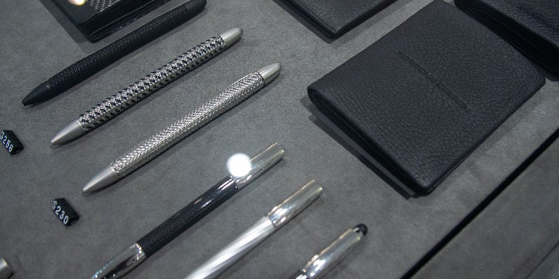 A display case with luxurious pens, including black and metallic woven textures, next to black leather wallets.