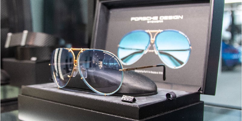 Porsche Design sunglasses with blue gradient lenses and gold frames are displayed on a stand inside a black box.